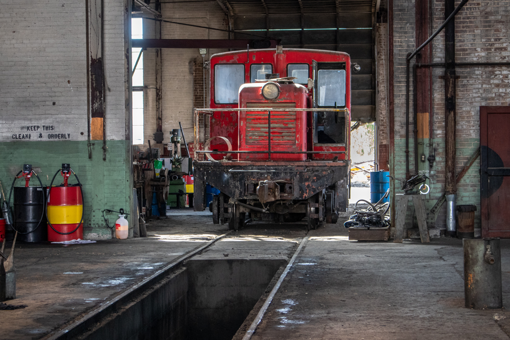 A small, red, locomotive parked inside an old brick enginehouse with an open inspection pit in the foreground