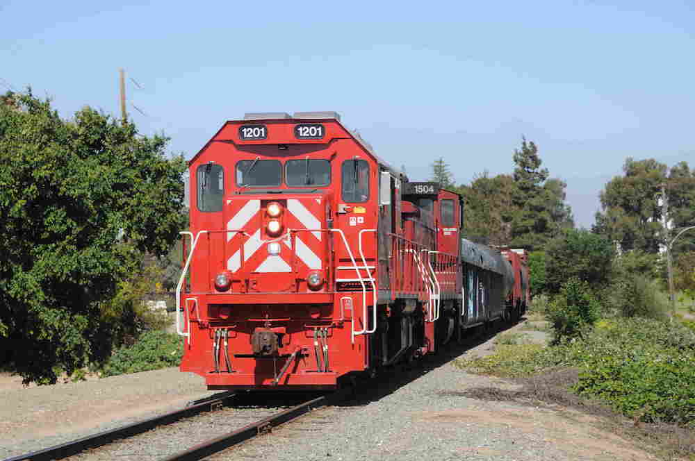 Two red locomotives pull a train down a rail line lined with small trees.