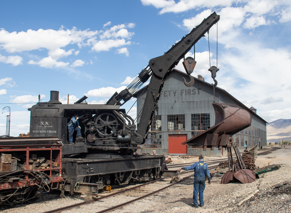 A black-painted steam wrecking crane lettered NN lifts a rust-colored snow plow blade