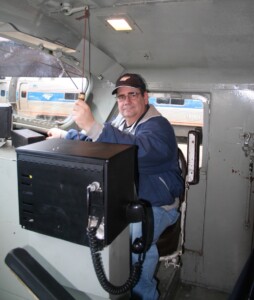 An interior image showing the inside of a locomotive control cab and a man in the engineer's seat