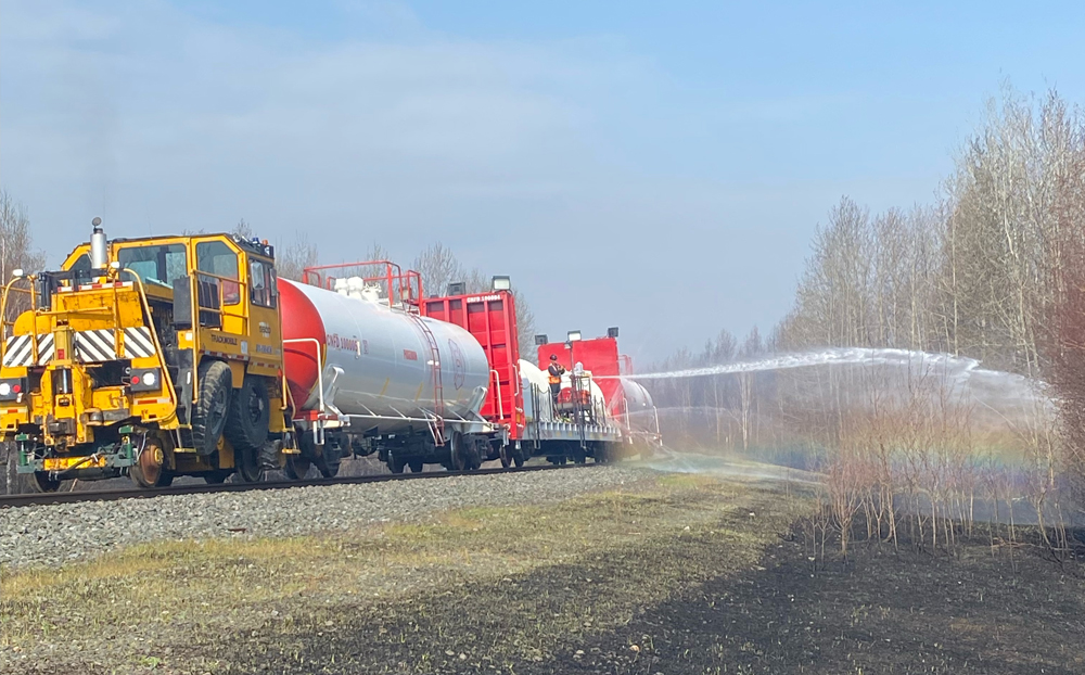 White tank cars, a flatcar with water cannons, and a trackmobile spray water along a railroad right-of-way.