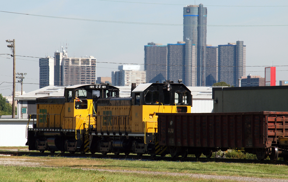 Two yellow end-cab switchers moving cars with city skyline in distance