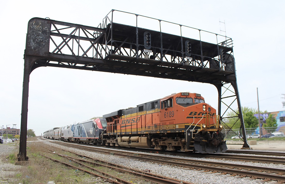 Passenger train led by orange freight locomotive passes under signal bridge