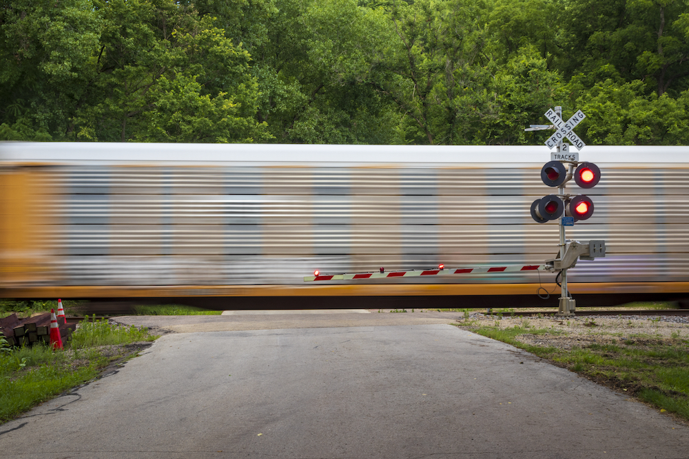 A side view of a train passing a rural grade crossing with railroad crossing sign, a lowered gate, and flashing red warning lights