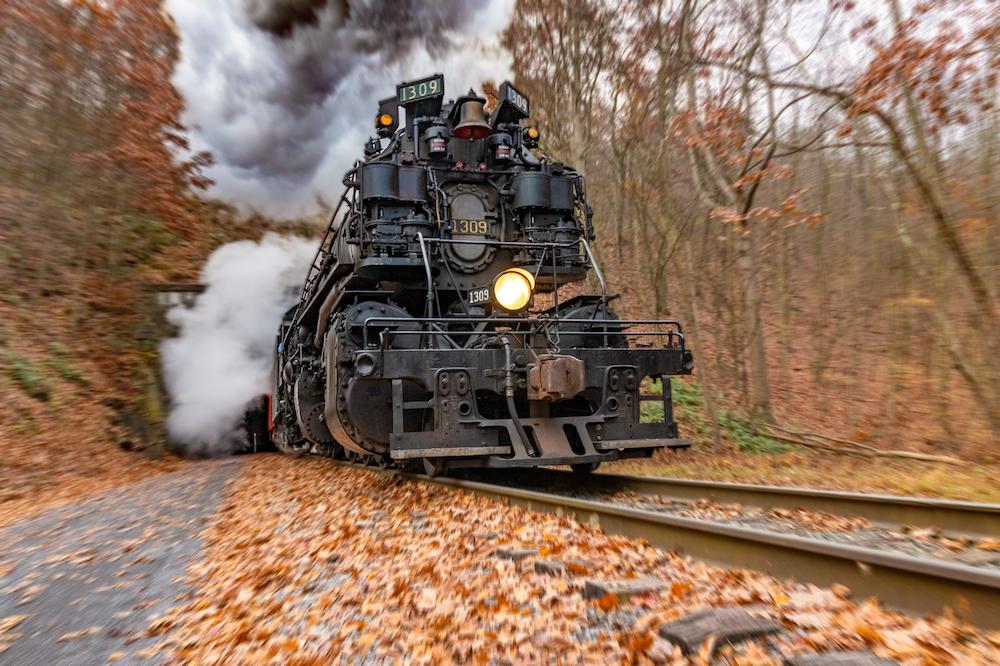 A large black steam locomotive, numbered 1309, emerges from a tunnel trailing a cloud of black smoke