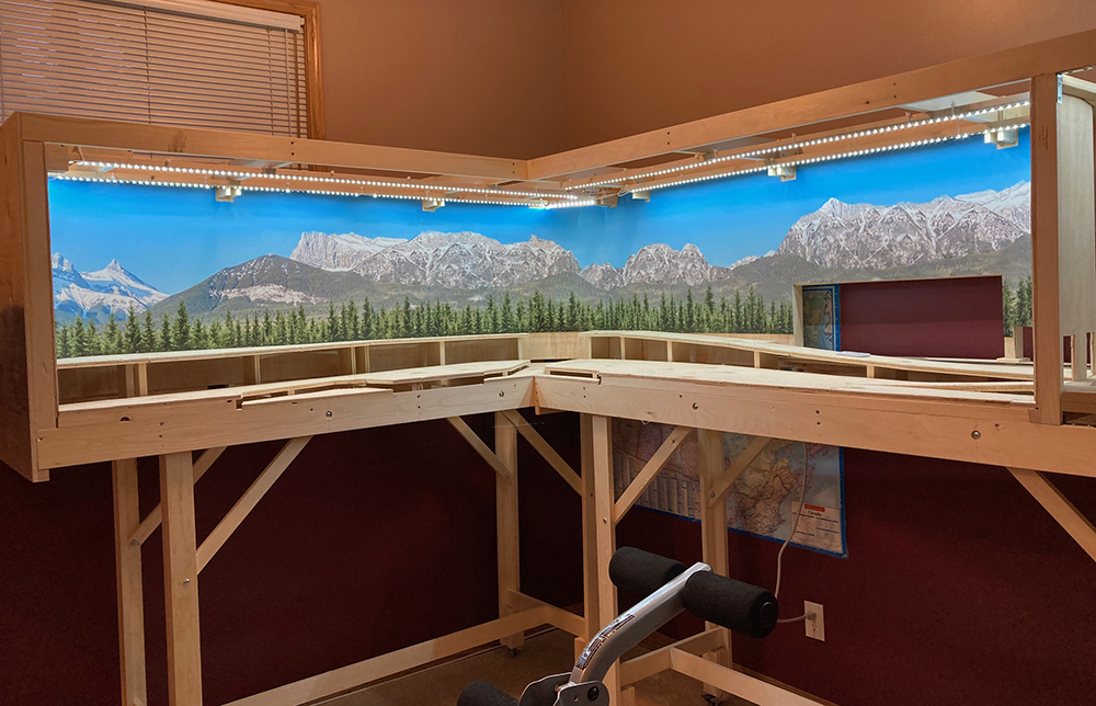 Jean Levasseur’s GP & Sons Lumber Co. in HO scale: Two tan wood rectangular boxes supported by bare wood legs. Inside the boxes is a mountain scene with blue sky, gray and white rocks, and green trees.
