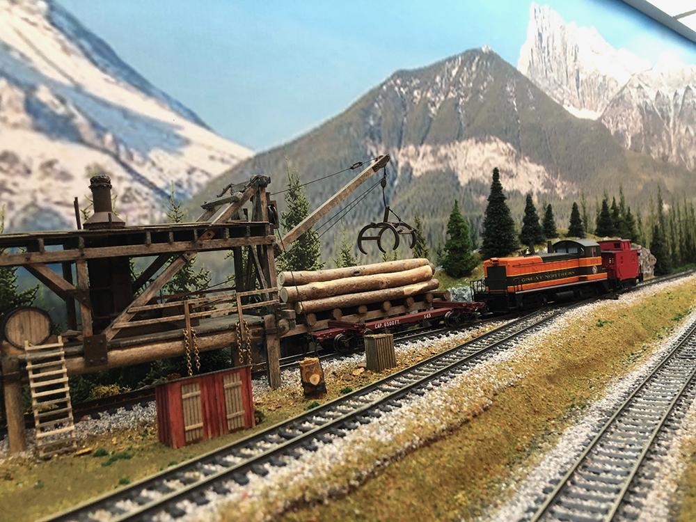 Model wood structure raised over train tracks with long arm and dark brown pincers for picking up logs