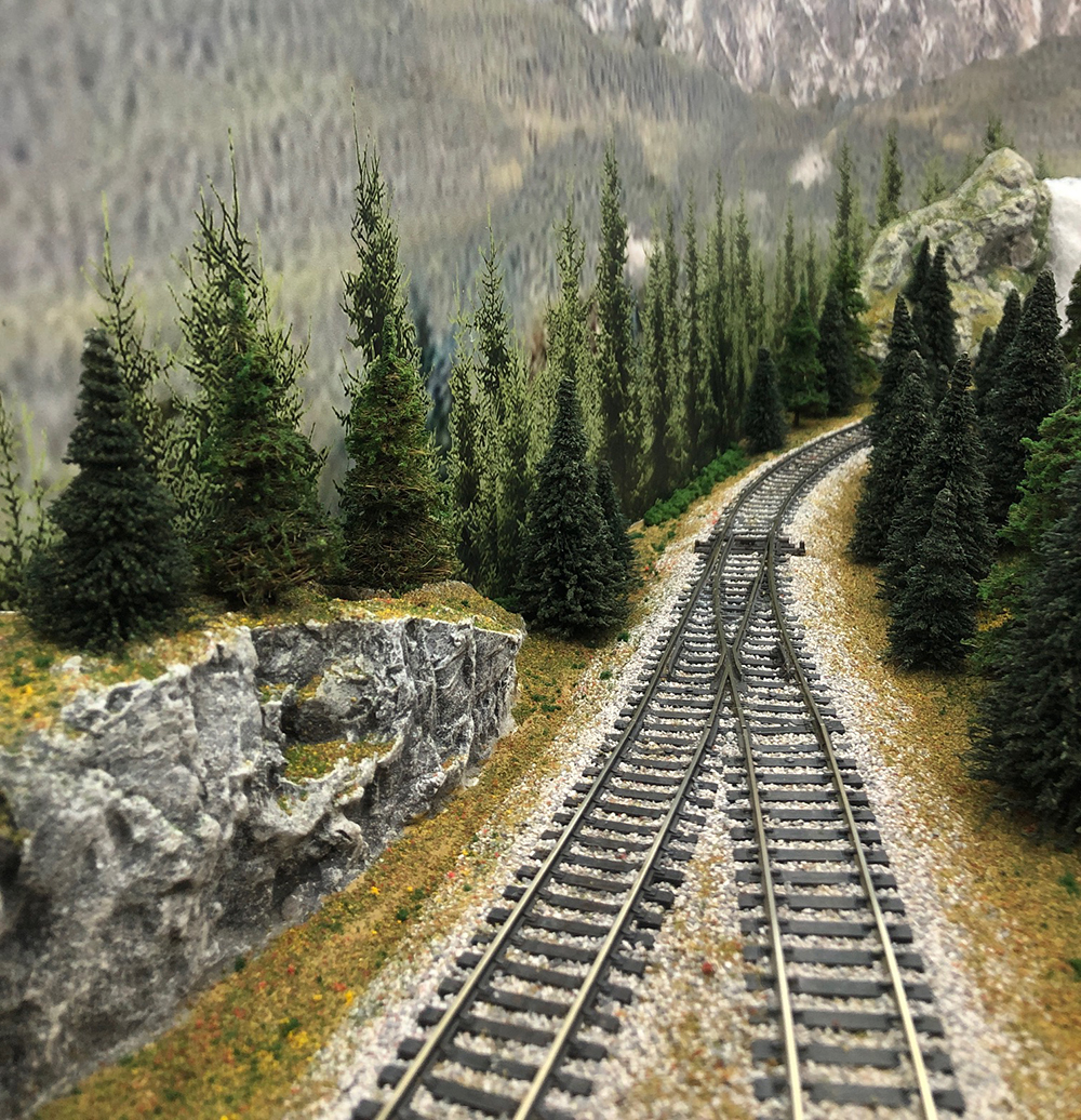 Jean Levasseur’s GP & Sons Lumber Co. in HO scale: Brown model railroad track with gray ballast passing through a gray model rock cut topped with green trees