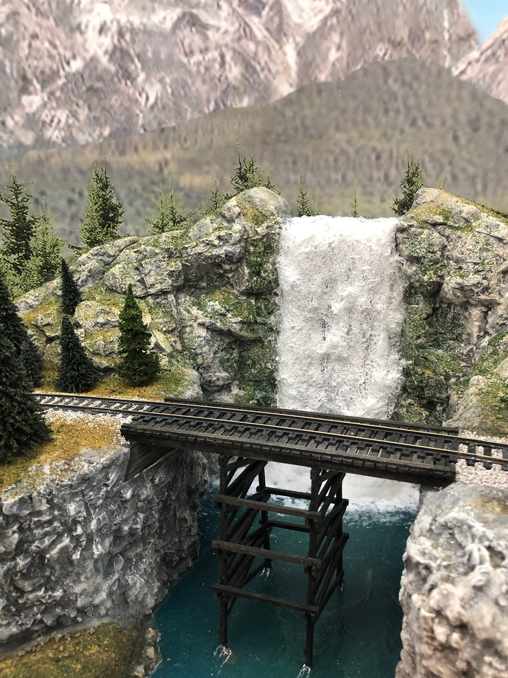 model waterfall with white water falling into a blue river that passes under a brown wooden railroad bridge