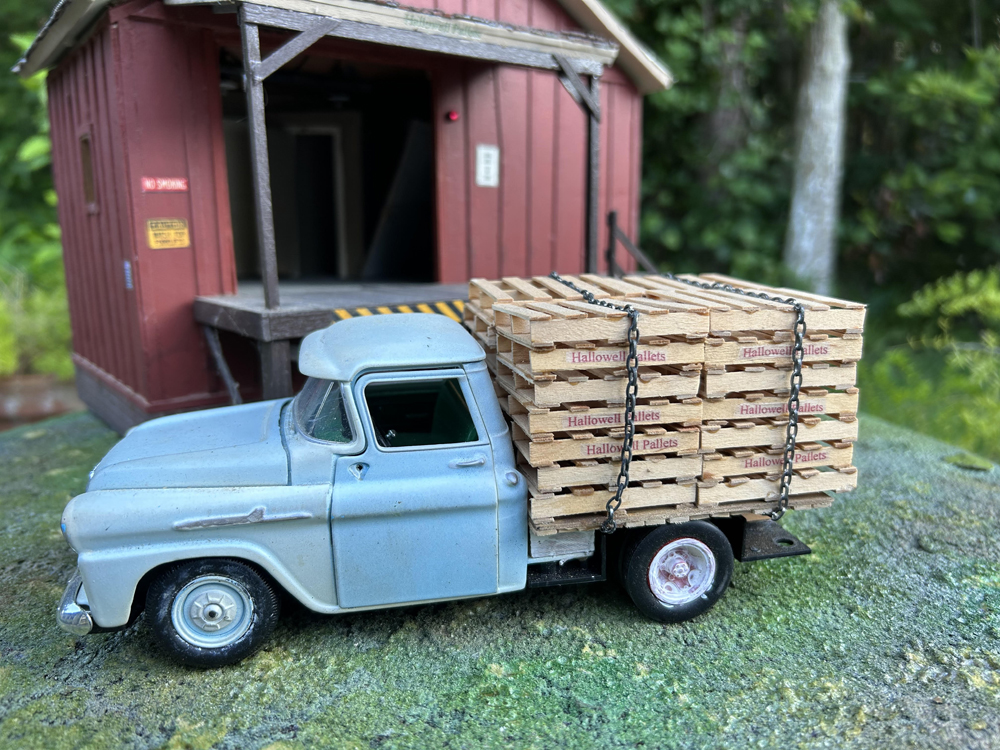 model blue truck with load of wood pallets in front of model structure