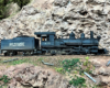 An outside-frame steam locomotive pulls a gondola past a rock wall