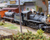 A small steam engine pulls a black tank car and orange refrigerated car past a brick freight house