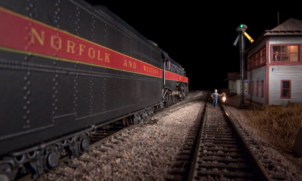 A towerman holds a lighted lantern aloft while a steam engine passes an interlocking tower at night