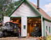A small steam engine emerges from a white-and-green wooden engine shed