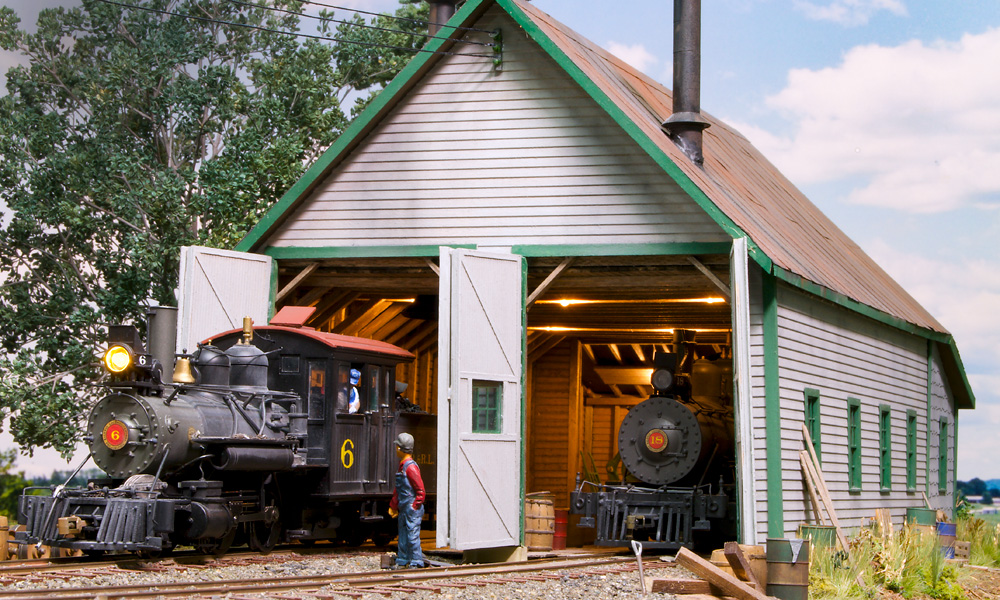 A small steam engine emerges from a white-and-green wooden engine shed