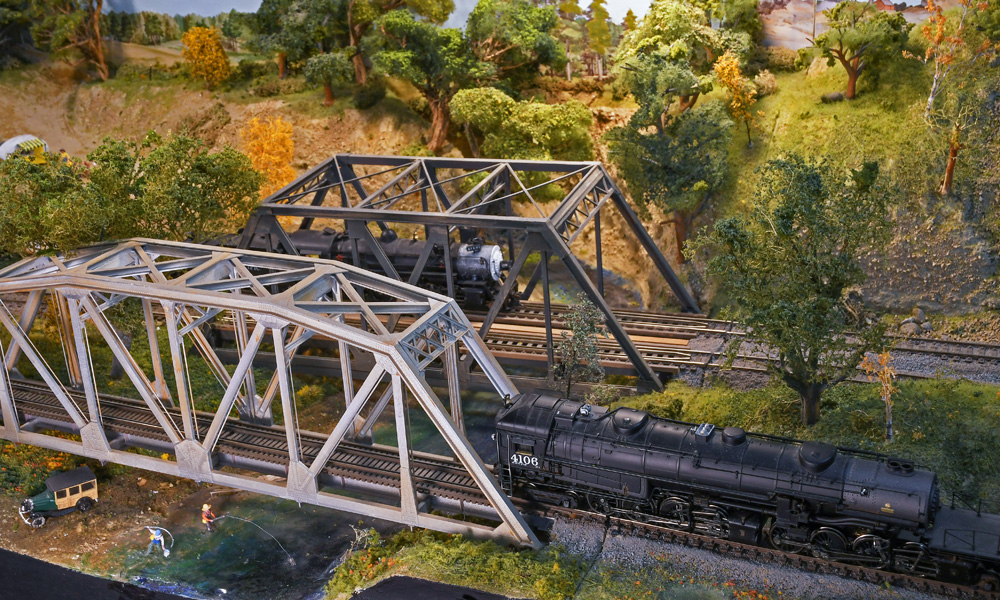 Two steam engines pass each other on two steel truss bridges that span the same creek