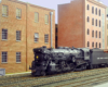 A black steam locomotive pulls a train past a row of brick industrial buildings