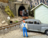 A short steam locomotive pulling gondolas emerges from a tunnel