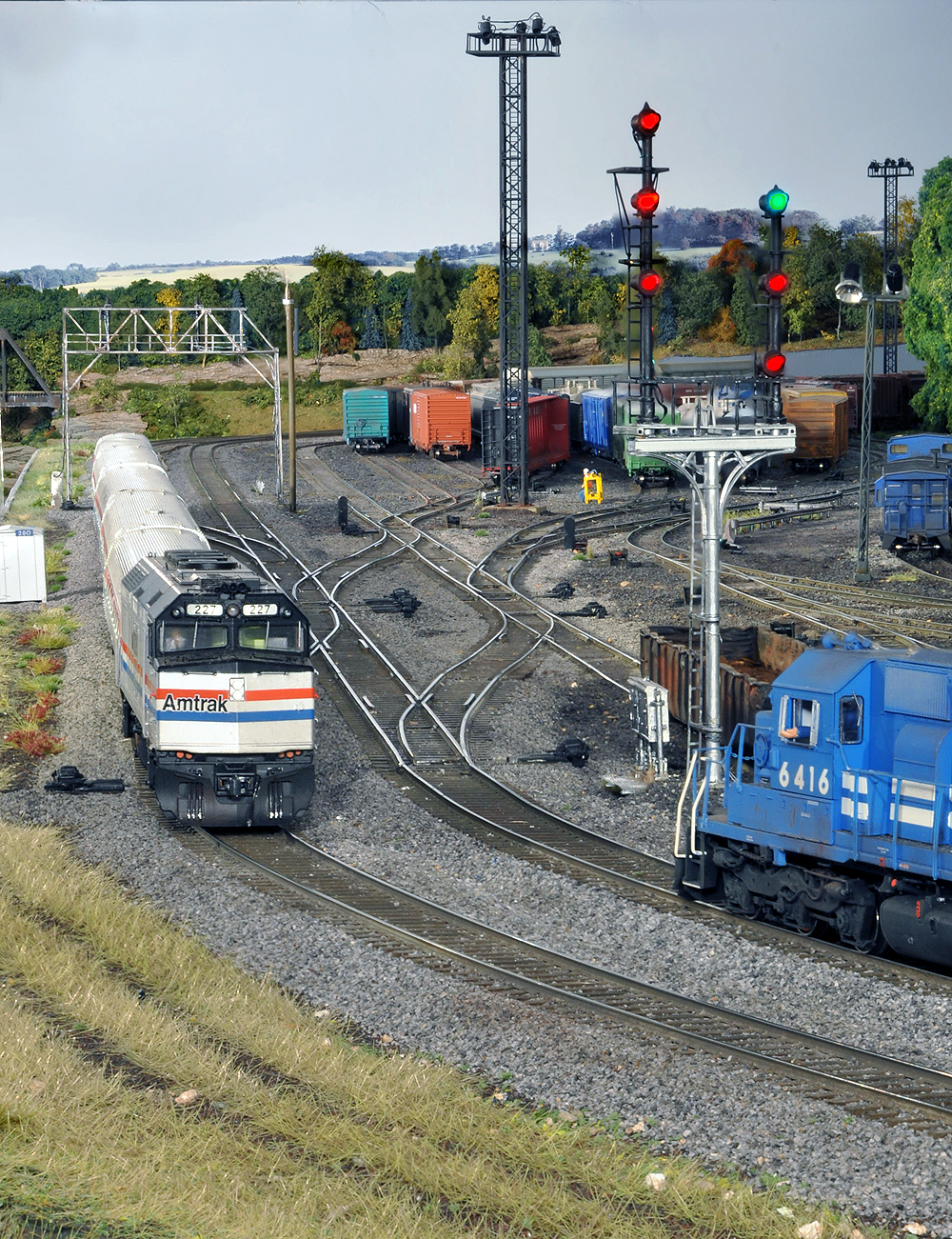 A silver Amtrak F40 meets a Conrail blue SD diesel as tracks branch off to the right into a rail yard.