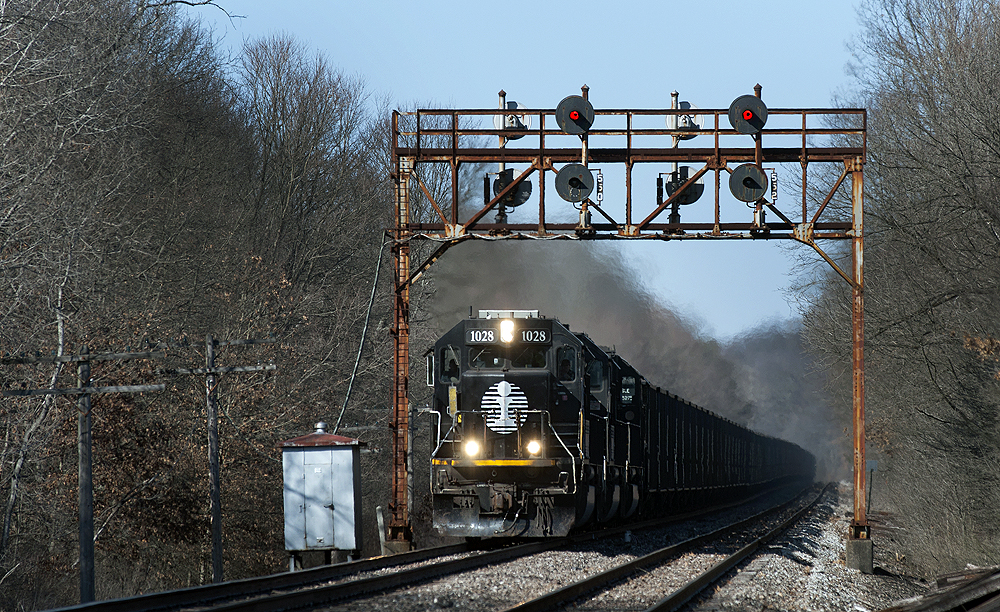 A rusted silver signal bridge carries searchlight signals across a double-track main line with a black EMD diesel pulling a freight train.