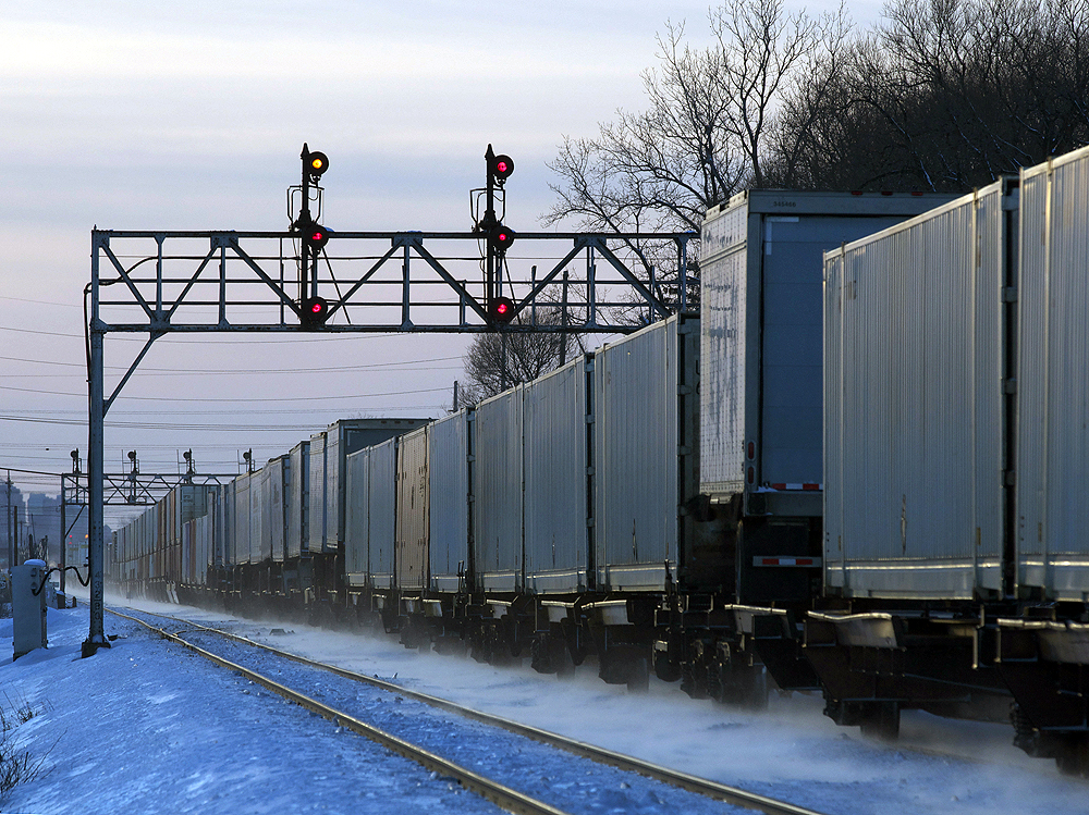 An intermodal train of containers and trailers on flatcars passes under a signal bridge on a clear winter day, kicking up snow as it goes.