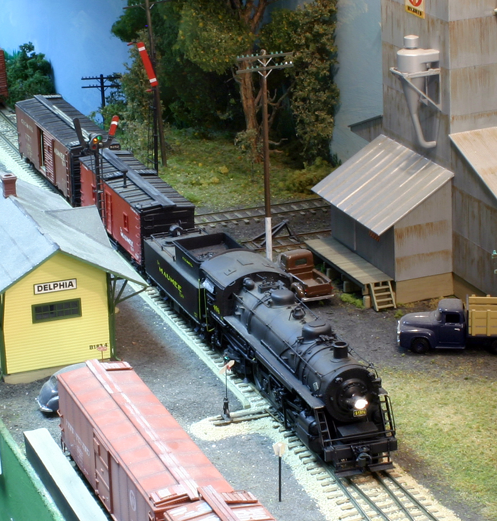 A steam locomotive works to spot two boxcars under a pair of semaphore signals’ vertical red blades.
