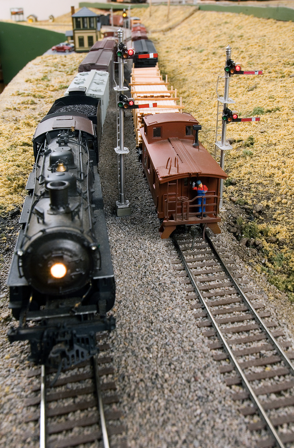 A freight trains led by a steam locomotive on the left is passed by a red caboose splitting a pair of semaphore signals on the siding to the right.