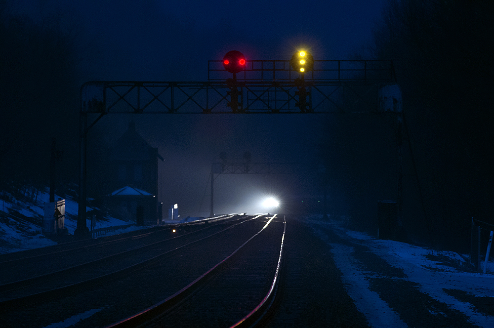 A headlight glows in the distance beneath a signal bridge with two red lights across a position light signal next to another showing a vertical line of pale yellow lights.