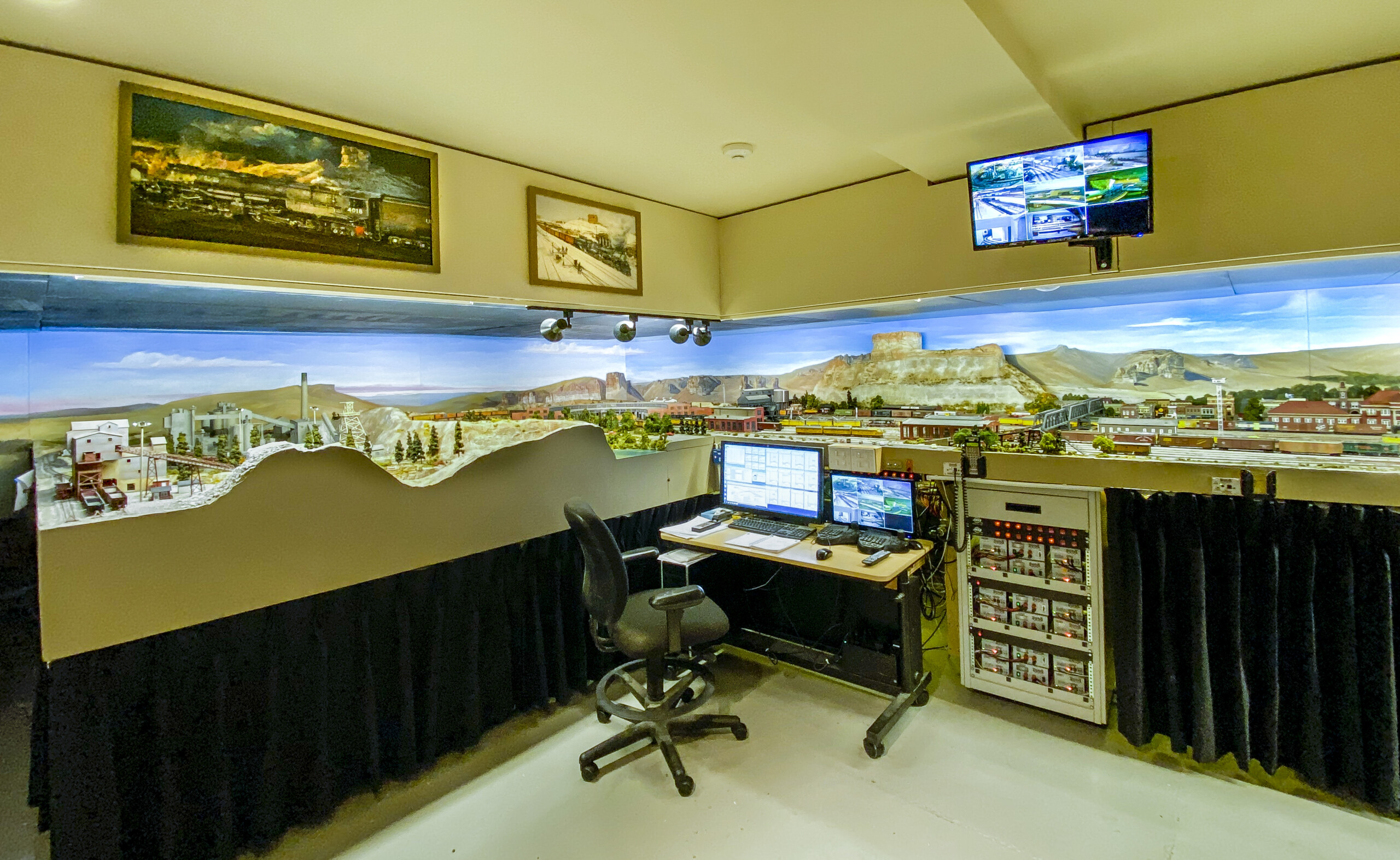 A desk with camera feeds is shown in front of the layout, with an additional monitor displaying camera feeds mounted above the layout.