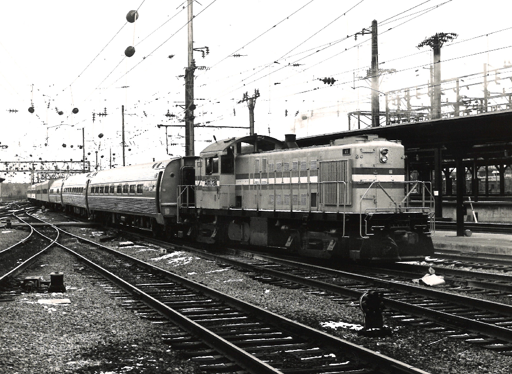 black and white photo of a diesel switch engine pulling passenger cars.