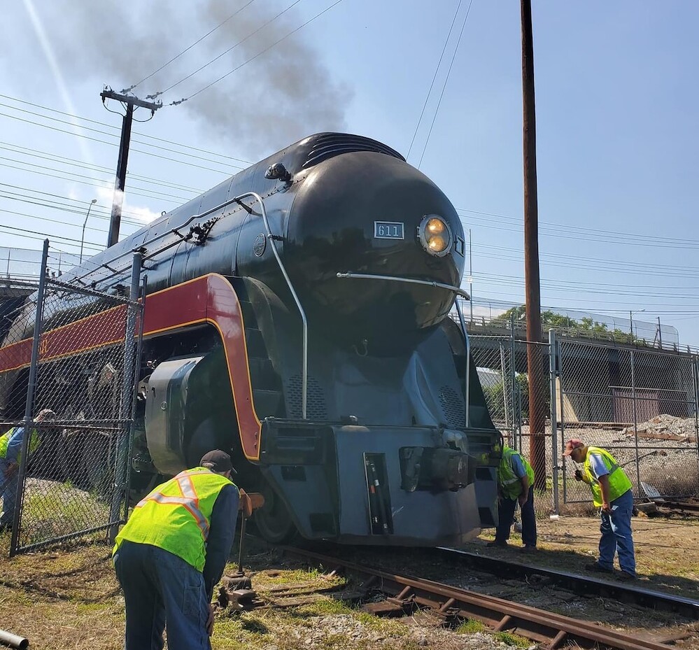 Steam locomotive pulls into yard while crew watches 