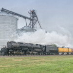 Large steam locomotive leads train past grain silo.
