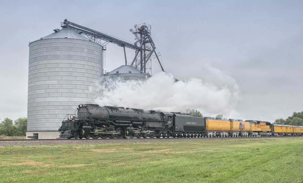 Large steam locomotive leads train past grain silo.