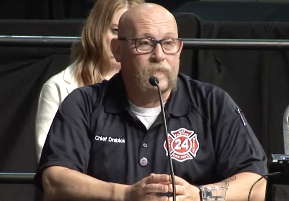 Man in black shirt with fire department logo speaks into microphone at hearing