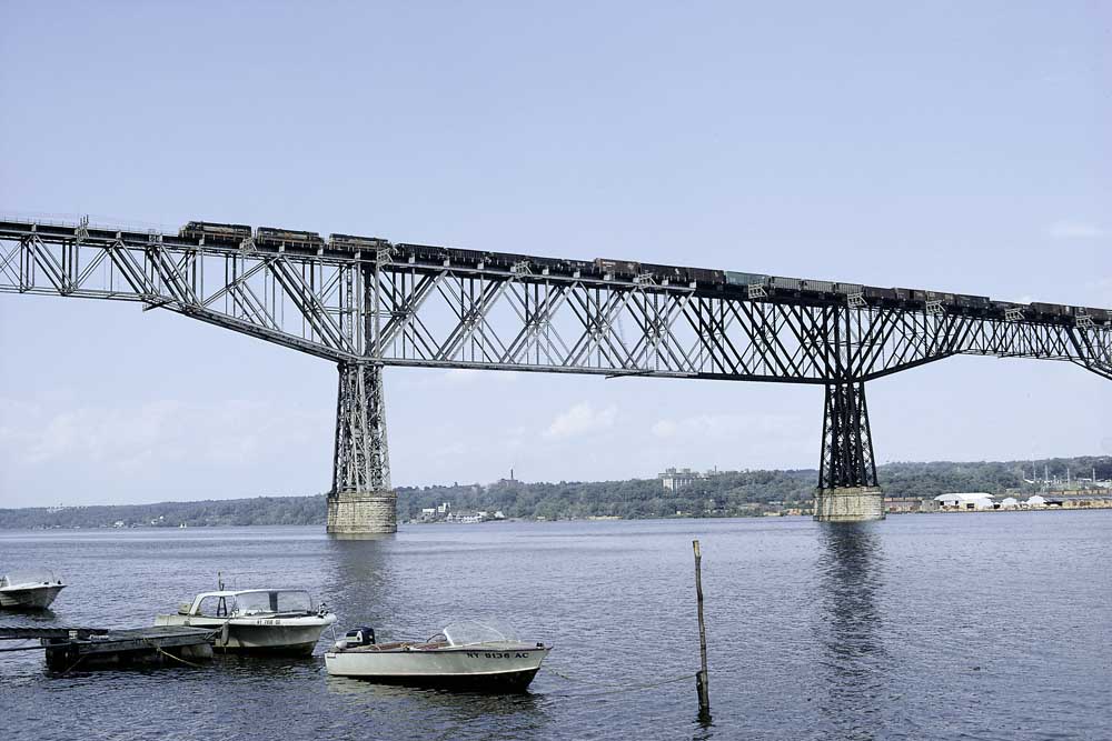 Three black diesel locomotives on tall bridge over water with boats in foreground