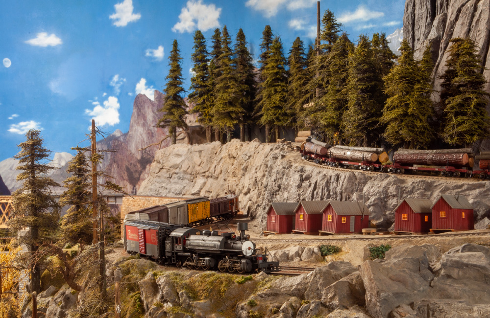 A manifest freight snakes past a cluster of logging cabins while a log train descends a steep grade on a cliff above