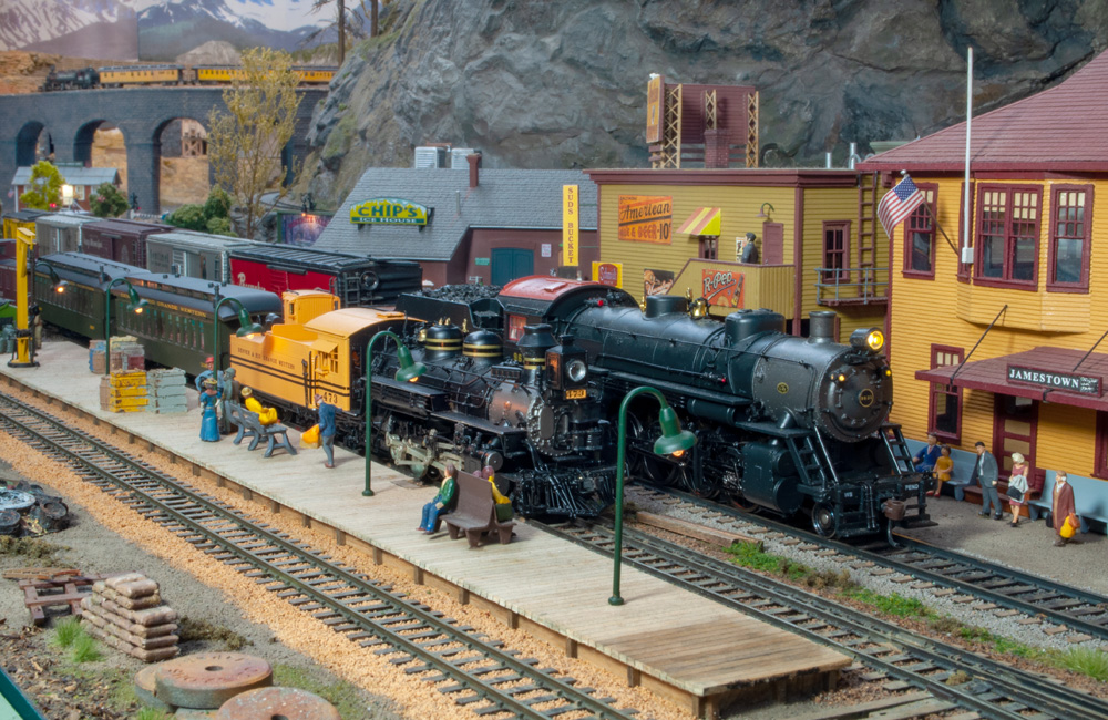 Steam-powered trains are seen in front of a yellow two-story station building