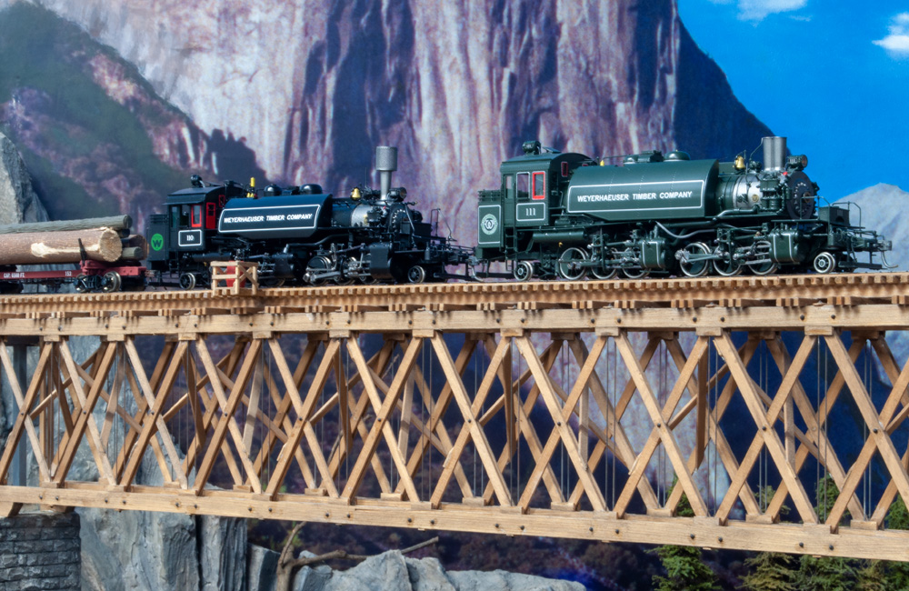 A black steam locomotive and a green one pull a log train across a wooden deck truss bridge