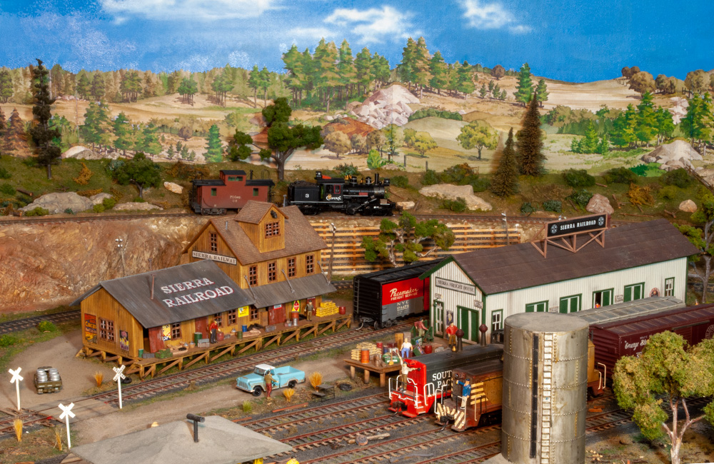 A small geared steam locomotive and caboose roll down the high line behind a freight yard