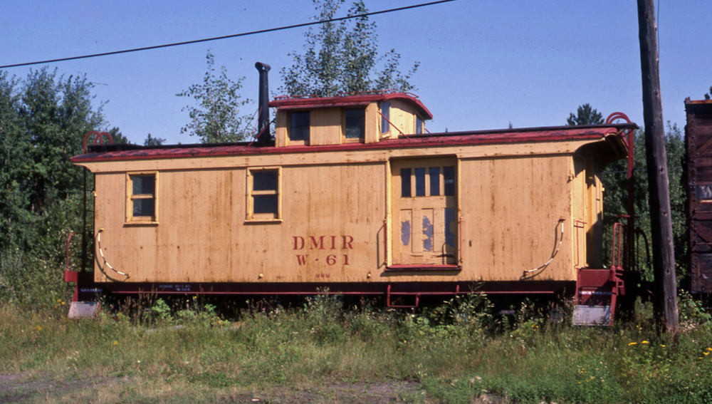 Color photo of wood caboose painted maroon and yellow with trees and utility pole in frame.