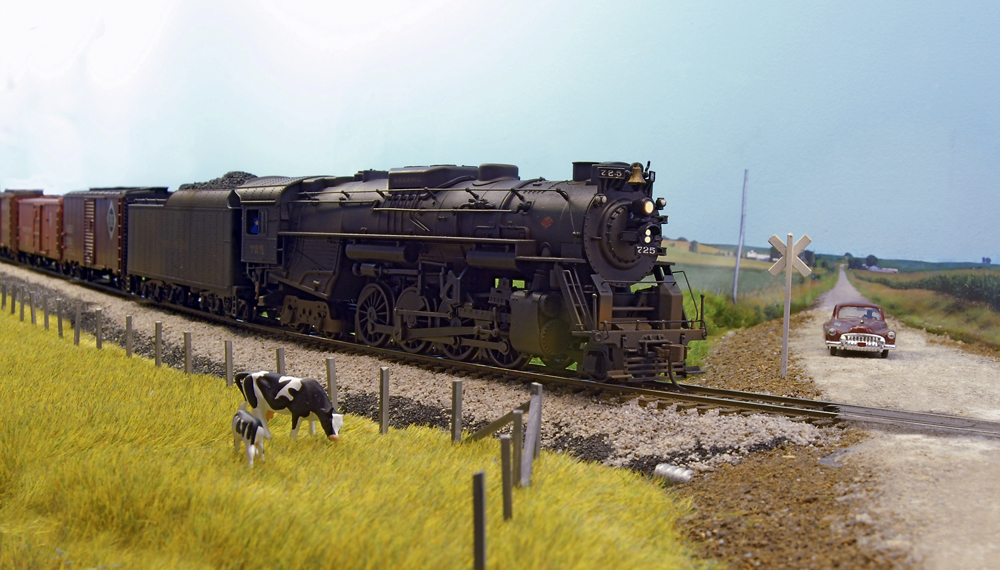 Color photo of steam engine with train approaching rural grade crossing.