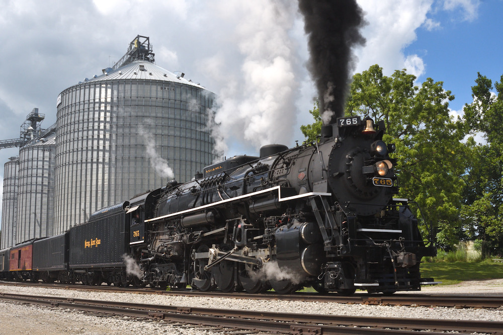 Steam locomotive pulls excursion train with grain elevator in the background.