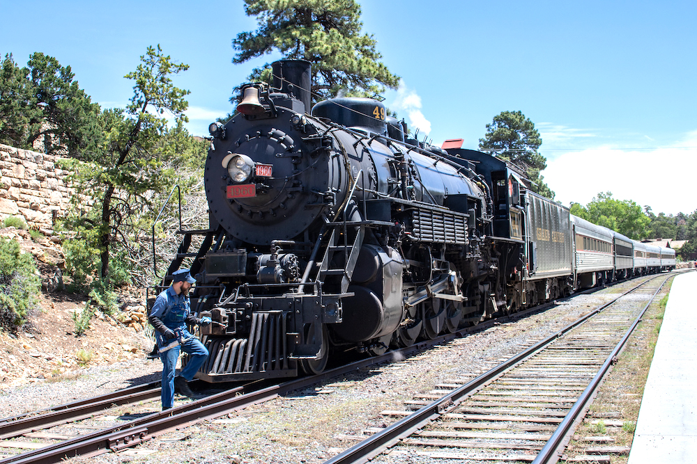 A crew member in blue coveralls checks the front of a parked black steam locomotive on a bright, sunny day.