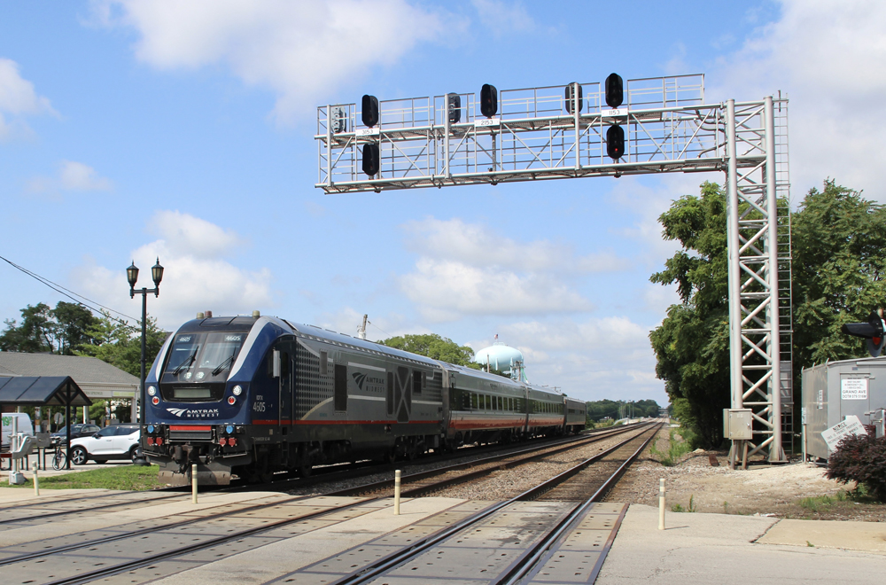 Passenger train with locomotive and three cars passes under signal bridge at grade crossing