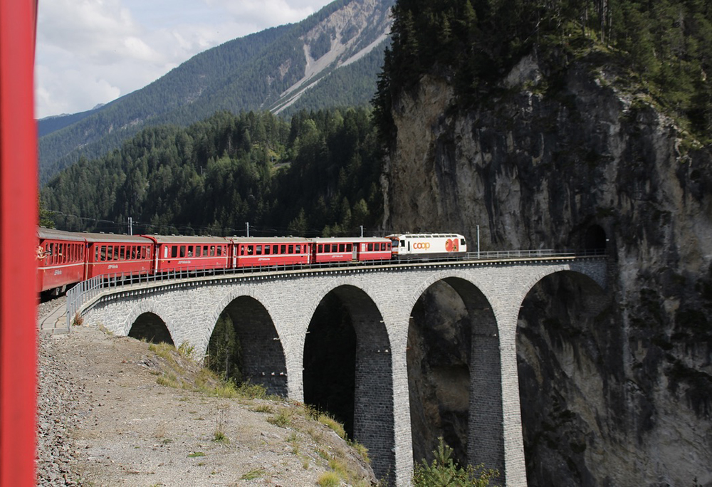 Train with red passenger cars on curving stone bridge leading to tunnel