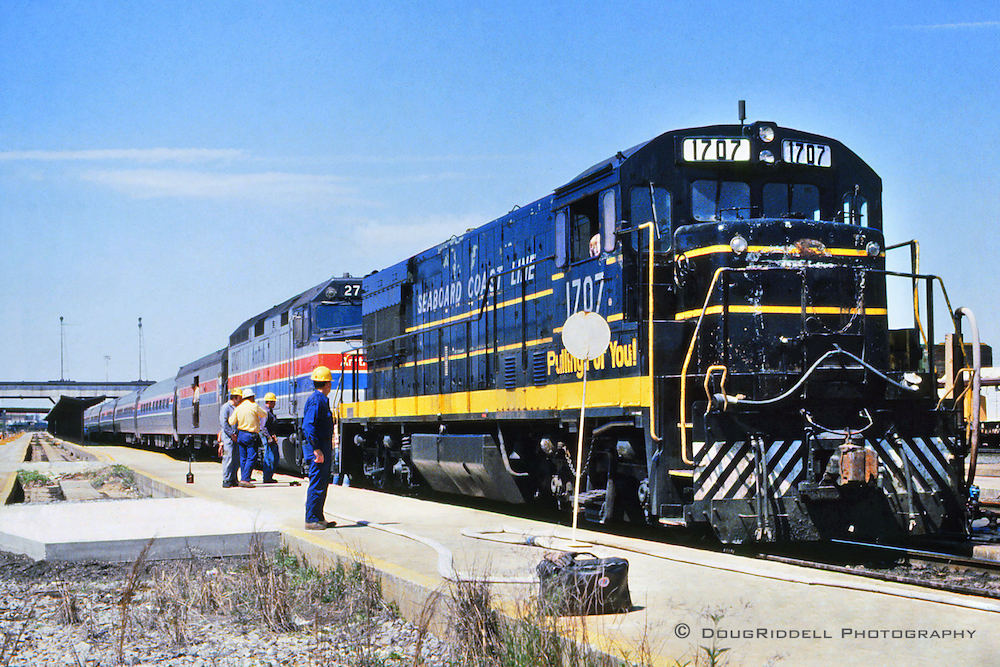 A dark-colored freight engine is coupled ahead of a silver Amtrak locomotive next to a concrete walkway.