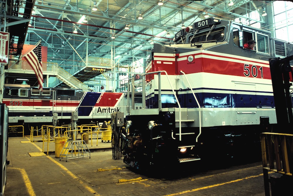 A gray locomotive with wide red, white, and blue stripes sits in a large and brightly lit factory building. A similar engine is in the background.