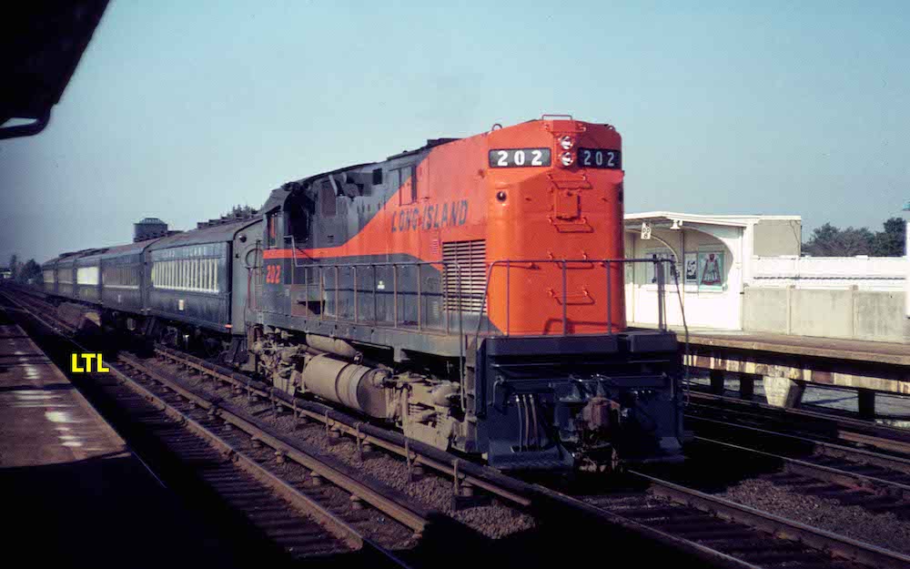 A commuter train powered by a diesel engine running long-hood-foward passes passenger platforms on either side of a triple-track mainline.