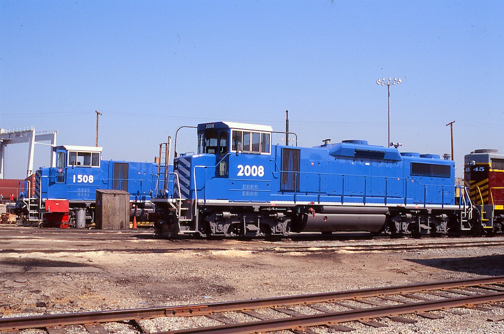 A pair of blue locomotives rest in a freight yard under a sunny sky. The locomotive have white roofs and white road numbers.