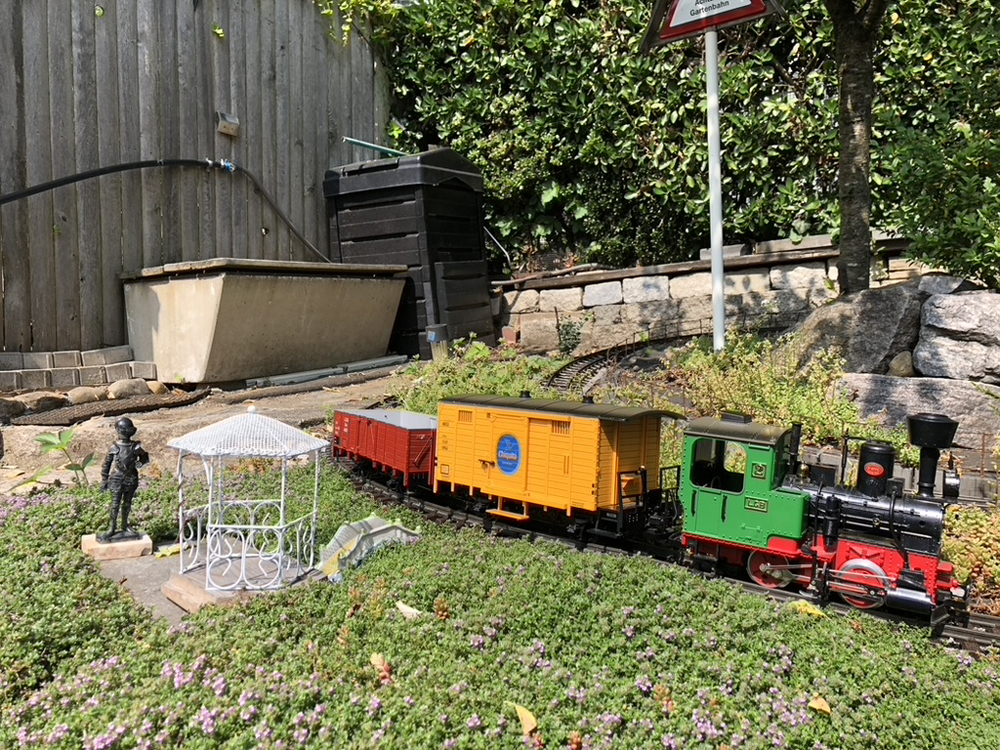 colorful model train on garden railway, with compost bin behind